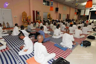 A seated group of devotees, mostly men in white clothes, sit in rows on striped red-and-blue floor mats in a hall with pink walls and orange hanging decorations. At the front, a golden throne-like seat and deity murti stand beside colourful figures, and a central figure sits on a raised seat facing the gathering. Water bottles and backpacks rest on the floor, and a large screen hangs at the right. The setting suggests a satsang discourse session.