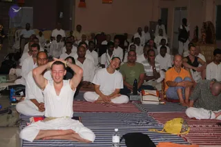 A large group of people sits on striped blue and red mats in a hall during a seated yoga session. In the foreground, a young man in white clothes sits cross-legged with both hands raised overhead, palms together, eyes closed. Beside him a man in a white kurta sits with hands folded, and a man in a green t-shirt and a man in a grey-and-white striped shirt sit nearby. A bald man in an orange shirt and blue shorts sits on a mat at right, and another man in a grey patterned shirt bows forward at far right. Behind them many men in white kurtas and dhotis sit in rows. Water bottles and bags lie on the mats, and a deity murti and decorative altar stand at upper right. No a person candidate is visible.