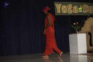 A photograph of a single man walking barefoot across a dark stage from left to right, head bowed. He wears a multicoloured turban with red and pink tones, an orange-red sleeveless garment with gold-trimmed edges wrapped as a shawl, and orange-red draped trousers with the same trim. A beaded necklace hangs at his chest. Behind him a dark blue curtain hangs beside a banner reading Yoga Sutras with a yoga figure graphic. On the right stands a white pedestal holding a small vase of red and white flowers, beside a golden armchair with dark wooden arms. The stage floor is glossy and reflects the figures.