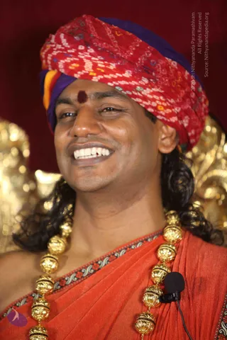 Close-up photograph of a smiling man in a red and purple bandhani turban with a red tilak on his forehead, long dark hair falling past his shoulders, wearing an orange-red shawl with a patterned border and heavy gold bead necklaces. A small black lavalier microphone is clipped to his garment. Gold ornate decorations blur in the background against a deep red backdrop.