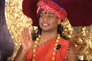 A man in an orange-saffron shawl with a red, yellow and purple turban and a red tilak mark on his forehead sits in front of an ornate gold-carved throne with a dark red backdrop. He wears a long gold bead necklace and raises his right hand in a blessing gesture while speaking. A small clip-on lapel microphone is attached to his shawl. Swamiji (probable): long dark hair, saffron garments, ceremonial gold beads and turban, appearance-based label only.