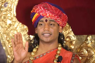 A man seated before an ornate gold-carved throne speaks while raising his right palm in a gesture of blessing or teaching. He wears a multicolored red, yellow and purple bandhani-style turban, a red-brown tilak on his forehead, long dark hair, an orange-red saffron shawl with a patterned border, and heavy ceremonial gold bead necklaces. A small clip microphone is pinned to his shawl. Behind him is a deep red backdrop.