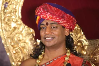 A close photograph of a man seated before an ornate gold throne with carved scrollwork and a deep red backdrop. He wears a red, purple and yellow bandhani-patterned turban, a red dot on his forehead, and long dark swept-back hair. He has a red-orange sannyasi-style wrap with a dark embroidered border and a heavy gold ceremonial necklace of beads. A small microphone is visible near his chest. He appears to be speaking, with a slight smile. Pictured in a seated discourse setting.