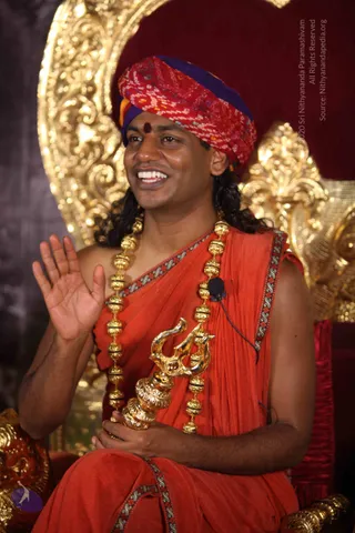 A smiling man seated on an ornate gold-carved throne against a deep red backdrop, waving his right hand. He wears a red and purple bandhani-patterned turban, a small red bindi, an orange-red shawl with a patterned border, and a long gold bead necklace. He holds a gold Om-shaped ceremonial object in his left hand, and a small clip-on lapel microphone is attached to his garment.