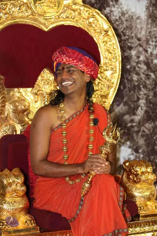 A smiling man seated on an ornate gold-and-red throne with carved gold lion-shaped armrests. He wears a red and purple patterned turban, an orange-saffron shawl with decorative border, long gold bead necklaces, and holds a gold trident-topped staff across his lap; a small lapel microphone is clipped to his shawl. A red tilak mark is on his forehead and long dark hair falls past his shoulders. Behind the throne is a patterned backdrop with a faded photographic print of trees.