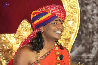 A smiling man sits before a gilded ornate throne with a red cushioned backrest. He wears a multicoloured bandhani turban in red, orange, yellow, green and purple, with a small red tilak on his forehead. His hair falls long and dark past his shoulders. He is bare-chested beneath a saffron-orange shawl with a embroidered border, and wears a long ceremonial gold necklace of beads and a lavaliere microphone clipped to the shawl. Appearance cues include the long hair, colourful turban, saffron clothing and gold necklace, consistent with the Swamiji (probable) label for this satsang photo. A gilded peetha-style backdrop and a stone-textured wall surround him.