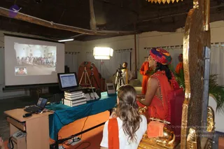 A seated figure in a saffron-orange sari with a multicolored turban sits on a golden ornate throne-like chair in a plain hall, facing a projection screen. The screen shows a crowd of people in a white-walled room and a small inset image. Beside the throne, a person in white clothing with an orange shawl sits with their back to the camera. A laptop, stacked books and a camera on a tripod stand on a teal-covered table, and a standing figure in orange robes is nearby under a studio light.