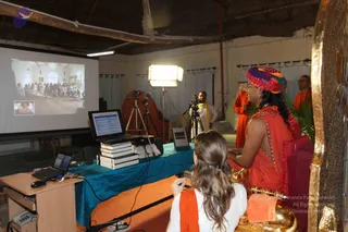 A photograph of an indoor satsang setting with a projection screen on the left showing a crowd in a hall. In the foreground, a woman with long light-brown hair in a white top and orange shawl sits facing the screen. Beside her, a person in a red-orange robe with a multicolored patterned turban sits on a golden throne-style seat with a red cushion, with palm ffronds at the side. Behind them, a man in white with an orange shawl stands near a camera on a tripod, and another person in orange robes stands at the right. A laptop, stacked books and a blue-draped table sit in the middle, with a bright studio light and white curtains behind.