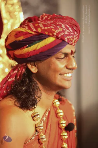 Close side-profile photograph of a smiling man wearing a multicoloured bandhani turban in red, yellow, purple and green, with a red tilak on his forehead. Long dark hair falls behind his shoulder and he wears an orange-red shawl with heavy gold ceremonial beads draped across his chest. A small black lapel microphone is clipped to the shawl. A blurred golden bokeh backdrop sits behind him.