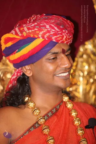 Close photograph of a smiling man in a red, purple, yellow and green bandhani-patterned turban, with a red tilak on his forehead. He wears a red shawl with a patterned border and a long necklace of large gold beads with red-and-black accents, and a small clip-on microphone is visible at lower right. A gold ornate chair stands behind him against a deep red backdrop.