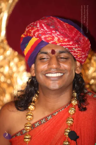 Close photograph of a smiling man wearing a red, purple and yellow bandhani-patterned turban and a red dot on his forehead. He has long dark hair and wears a red garment edged with embroidered border and a necklace of gold beaded spheres; a small lapel microphone is clipped to his clothing. A glowing golden background sits behind him against a deep red backdrop. The man is visible from the head to the chest and appears to be seated before a gilded throne-like setting.