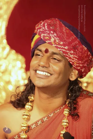 Close photo of a smiling man wearing a red, purple and yellow bandhani-patterned turban with a red bindi on his forehead. He has long dark wavy hair falling past his shoulders and wears a red-orange shawl with a patterned border and heavy ornate gold bead necklaces. A small microphone is visible at the bottom edge, and a warm gold bokeh background sits behind him against a deep red backdrop.
