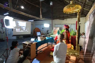 A photo of a satsang room with a thatched roof and white walls. A person in an orange robe and a multicoloured turban stands near a gold ornate palanquin-style seat with a gold canopy, facing a projector screen. A bald man in white clothing kneels in front of the seat. Two women in white clothes with orange shawls sit at a wooden table with laptops, one operating a laptop. Stacked books, studio lights and a camera on a tripod are visible, along with a framed painting, a flag and potted palms.