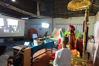 A photograph of a satsang room with a thatched roof and white walls. On the right, a robed figure in saffron-orange clothing with a multicoloured turban stands beside an ornate gold canopy and throne-like seat with red cushions, looking up toward the screen. A shaved-headed man in white clothes sits on the floor in the foreground, facing the throne. A large projector screen on the left shows a video, and a laptop and stacked books sit on a teal-covered table. A camera on a tripod and studio lights are set up, and a framed painting hangs on the wall. A man in white sits at the far right edge, and a person in orange works a laptop at the lower left. A green sofa and potted palm complete the setting.