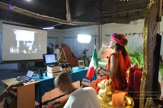 A satsang discourse in a bright hall with a bamboo-beamed ceiling and white walls. On the right, a figure in orange robes and a multicoloured red, purple and green turban sits on an ornate red-cushioned throne with gold lion-shaped armrests, gesturing with one hand while speaking. A projector screen on the left shows a live feed of a seated figure in orange. In front, a laptop, a blue-draped table stacked with books, a small clock and an Italian-style tricolour flag. Foreground: a shaven-headed person in white clothing seen from behind, a man in a yellow top at the back operating a camera on a tripod, and additional lights and cameras. Potted palms stand at the right edge.