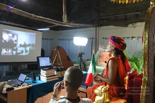 A photograph of a satsang talk in a simple hall with a corrugated roof. A seated person in saffron-orange robes and a multicoloured turban sits on a gold-trimmed throne-like seat with a golden lion figure, holding a small object and looking toward a projection screen on the left. The screen shows a slide with a small inset photo of a similarly robed figure. A man with a shaved head in white clothes and a rudraksha mala holds a camera in the foreground. An Italian-style tricolour flag stands beside the speaker, and a laptop with stacked books sits on a blue-draped table. Studio lights and a tripod-mounted camera are set up behind. a person: the seated speaker wears saffron robes, a colourful turban and a rudraksha mala, and is seated on a throne. The foreground photographer is a separate person.