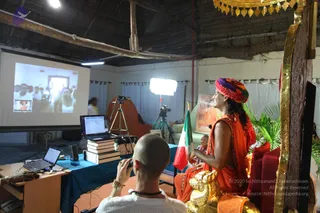 A photograph of a satsang session in a thatched-roof hall. On the right, a man in orange robes and a multicoloured red, yellow and blue turban sits on an ornate red and gold throne, with a golden lion figure at its base and a gold canopy above. A saffron-clad figure with a shaved head sits in front of a tripod-mounted camera. Behind them, a projector screen shows a crowd of people. A shaved-headed man in white with a rudraksha mala operates a camera at the front. Other people sit on a table draped in teal cloth beside a laptop and stacked books, and an Indian flag stands nearby. Potted palms and a framed picture are visible at the side of the room.