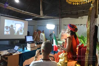 A photograph of an indoor satsang hall with a corrugated roof and white curtains. A person in a saffron robe and a multicoloured pink, orange and green turban sits on an ornate golden-and-red throne with a golden lion-shaped base, holding up a folded Indian tricolour flag and gesturing toward a projection screen. The screen shows a small group of people in white. Nearby, a man in white clothes with a yellow shawl stands, a shaved-headed man in white in the foreground photographs with a camera, and a laptop, stacked books and a tripod-mounted video camera sit on a blue-draped table. A framed portrait of a saffron-clad figure hangs on the wall beside a tall potted plant. Swamiji (probable): the seated figure wears a multicoloured turban, saffron robe and a throne, with long dark hair visible. The overlay reads 2020 Sri Nithyananda Paramashivam