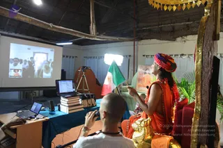 A photo inside a simple open-sided hall with a thatched ceiling and white curtains. A figure with long dark hair, a multicoloured patterned turban and a saffron-orange robe sits on a red and gold throne-like seat, holding up a green-white-red flag and looking upward. Beside the seat a large gilded lion-form ornament stands. Behind them is a framed portrait of a saffron-clad figure. In the foreground a bald man in a white shirt with a beaded necklace holds a camera toward the scene, and a laptop, stacked books and a tripod sit on a blue-draped table. A projection screen shows a group of people in a room.
