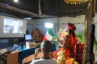 A seated figure in saffron-orange robes and a multicoloured turban sits on an ornate red and gold throne in a thatched-roof hall, holding a small Italian-style tricolour flag aloft while turning toward a projection screen. Beside him a shaved-headed man in white with beaded necklaces films with a camera on a tripod-mounted rig, and a third person works at a laptop on a table covered with stacked books. The screen shows a crowd and a small inset image. A framed picture of a deity hangs on the wall with potted palms nearby. The figure's appearance includes a multicoloured turban, saffron robes and a golden ceremonial necklace, consistent with Swamiji (probable) as the seated speaker on the throne.