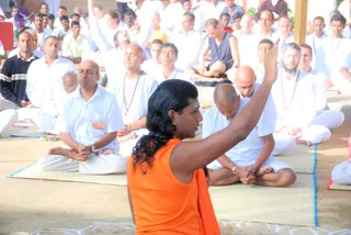 A photograph of an outdoor satsang where a figure in an orange sleeveless top and orange shawl sits cross-legged on a mat, long dark hair swept back, one arm raised with the hand open as if addressing the seated audience. Around them, many people in white clothing sit on mats in meditative poses, some with eyes closed, hands folded, and some wearing beaded malas. A bald man in white in the foreground bends forward with hands clasped. Hair and orange clothing of the speaker distinguish them from the white-clad audience. The setting is a sandy ground with a blue-edged mat and a red wall in the background.