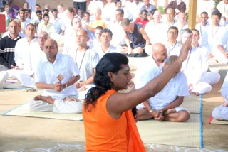 Outdoor satsang on a sandy ground with a large seated gathering dressed mostly in white. In the foreground, a long-haired man in an orange sleeveless top and orange shawl sits facing the crowd, his right arm raised with the hand open in a teaching gesture. Behind him, men in white kurtas and dhotis sit cross-legged on thin mats with blue borders, some with hands folded in prayer, and a man in a dark sleeveless top sits among them. Rudraksha malas hang on several chests.