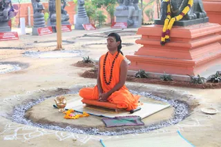A man sits cross-legged in meditation on a raised wooden seat atop a mat in an open outdoor courtyard. He wears an orange saffron robe, a long rudraksha mala around his neck, and a vibhuti/kumkum mark on his forehead, with his hands resting in his lap and eyes closed. Around him a circle of white rangoli-style pattern and grey ash-like powder marks the ground, with a brass kalasha/pot, a small offering of orange and yellow marigold garlands, and a small brass plate with a pink offering nearby. Behind him a large orange pedestal holds a dark stone deity murti draped in yellow and red flower garlands, and rows of grey stone statues and red placards stand in the background.