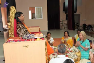 A photograph of a padapuja ceremony in a hall with beige walls, a window and dark red curtains. A man with long black hair, wearing a leopard-print shawl over a saffron garment and a saffron-orange cloth draped over his shoulder, sits on an ornate gold-carved throne with a red cushion, eyes closed. On a red-cushioned table before him are apples, sweets and a tray of offerings. Seated devotees in front of him include a woman in a yellow sari with hands pressed together in prayer, a woman in a teal salwar kameez with hands folded, a woman in white and yellow, and a man in white with a red-bead garland. Brass plates holding pink flowers, red fruit and small lamps sit on a gold cloth. Partially visible people sit at the bottom edge of the frame.