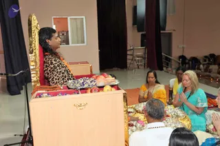 A photograph taken inside a plain hall with cream walls and dark curtains. A long-haired figure in a leopard-print top and an orange-and-rudraksha garland sits on an ornate gold-framed throne with red upholstery, speaking into a microphone on a stand. In front of the throne is a wooden table holding a tray of apples and wrapped sweets. Seated on the floor below, a woman in a yellow sari and a woman in a turquoise salwar kameez sit with palms pressed together in namaste, while a man in white clothes sits with a brass plate of offerings before him. Several other attendees are partially visible at the lower edge, and folding chairs line the far side of the room.