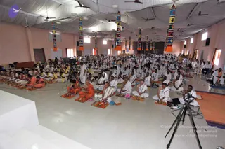 A wide view of a large hall during a seated puja gathering. Hundreds of people in white ritual clothes sit cross-legged on the floor in rows, many with small brass plates and oil lamps set on orange-and-red cloth mats in front of them. Several monks in saffron and orange robes sit among them, and a few cameras on tripods film the scene at the front. Tall cloth pillars in bright patterned blue, yellow and orange hang from a white draped ceiling, with ceiling fans overhead and a raised white platform at the left edge.