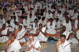 A large seated congregation of men in white ritual cloth with red-bordered dhotis and white shawls performs a pada puja-style ceremony on a glossy floor. Many have palms pressed together in prayer, and a few have white and orange sandalwood or ash marks on the forehead and arms. Small brass lamps, brass plates with fruit and flowers, and small silver-rimmed bowls sit on orange and yellow mats in front of them. Rudraksha malas hang around their necks. Behind them, rows of seated devotees fill the hall, some in orange or saffron clothing. No individual appears to be a clearly identifiable a person candidate.