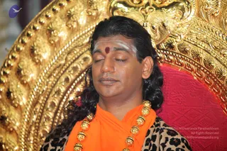 A photograph of a man with long dark swept-back hair, eyes closed, wearing a white vibhuti line and a red kumkum dot on his forehead. He wears an orange saffron shawl over a leopard-print garment and a heavy gold rudraksha-style bead necklace. He sits before an ornate gold carved throne with a red cushioned backrest. The figure appears to be Swamiji (probable) based on the long swept-back hair, saffron clothing, gold necklace and seated throne, though this is an appearance-based label only.