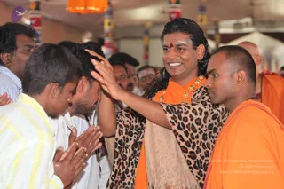 A photograph of a crowd at an indoor event. A man with long swept-back dark hair, a red bindi and white tilak, wearing a saffron-orange robe, a leopard-print shawl and a gold necklace of large ornaments, smiles while placing his raised hand on the head of a devotee bowing in front of him. Devotees in white shirts press their palms together in anjali. On the right, a shaved-headed attendant in orange robes stands close by, and more people in orange and white fill the background beneath colourful hanging decorations.
