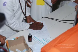 A photograph of a man in white clothing and a rudraksha mala, whose hand rests on a sheet of handwritten paper on a white table. Other people in white stand behind him, and a corner of an orange garment is visible at the right edge. On the table are a small brown glass bottle with a metal cap and a row of blue-capped small vials in a cardboard box, with a black cable running along the table. No one visible is identifiable as the a person, and no robed central figure is shown.