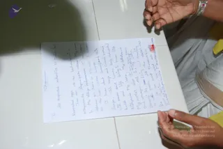 A handwritten letter in blue ink lies on a white tiled surface, with the top of the page reading a salutation to respected authorities of Indian law. Two hands are visible at the edges of the frame, one at upper right with a silver bracelet and one at lower right wearing a yellow garment, appearing to hold or steady the page. A red ink mark or seal appears near the upper right corner of the letter. Shadows fall across the upper left of the tiles. The letter is a personal handwritten statement with several paragraphs of cursive text.