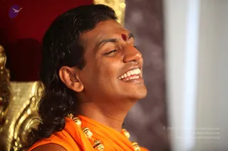 Close profile photo of a smiling man with long, swept-back dark hair and a red kumkum mark on his forehead. He wears an orange saffron garment and a gold-toned ceremonial necklace with rudraksha-style beads, seated in front of a gold ornate chair against a red and grey backdrop. Cues are distinctive: long hair, saffron clothing, rudraksha-style beads and gold ceremonial ornaments, so this is a Swamiji (probable) in person during a satsang session.