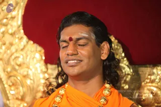 Close-up photo of a man with long dark wavy hair, eyes closed and a faint smile, seated in front of an ornate gold-carved throne against a deep red backdrop. He wears saffron-orange ceremonial clothing, a gold multi-bead necklace, and a red kumkum dot with white vibhuti marks on his forehead. Swamiji (probable): the saffron clothing, swept-back long hair, ceremonial gold necklace and throne setting are consistent with the page's Swamiji label, so the appearance-based candidate is physically present.