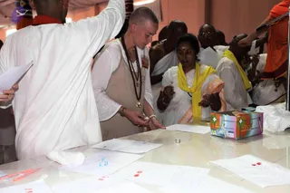 A photograph of a registration or ritual-preparation table at an indoor hall. A woman with a shaved head wears a beige sleeveless tunic over a white top, with a rudraksha mala and a pendant, and sorts papers on the table. Beside her a woman in a white sari with a yellow shawl and a red bindi holds a small object in her hands. Behind them several men in white and orange robes sit or stand. On the table are scattered sheets of paper with red markings, small blue vials, cotton wool, pens and a colourful Ball pens box. A man in a white kurta on the left raises one arm.