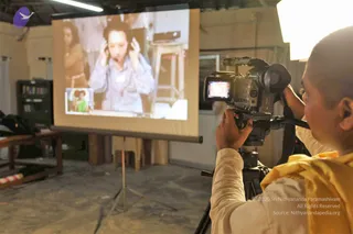 A cameraman in a cream shirt and yellow shawl films a projection screen with his handheld video camera on a tripod in a bare, unfinished room. The screen shows a woman in a light blue shirt with headphones on, and a small inset picture of a child in a green top. A black speaker stand and wooden furniture sit at the left, with a bright light fixture at the upper right.