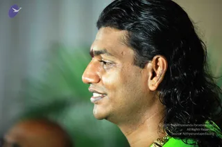 Close side-profile photograph of a man with long, swept-back black wavy hair, a red tilak mark on his forehead, and a green garment with a gold necklace at the bottom edge of the frame. He faces left and appears to be speaking or smiling, with a blurred green plant and grey background behind him.