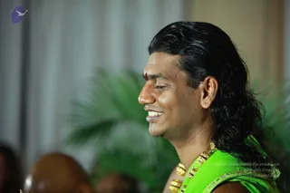 Side-profile photograph of a smiling man with long, swept-back dark wavy hair and a red tilak mark on his forehead. He wears a bright green silk garment with gold-trimmed border and a heavy gold ceremonial necklace with gold beads. Blurred green palm fronds and a pale curtain form the background, and the blurred head of another person is visible at lower left. a person: the long swept-back hair, gold ceremonial necklace and ornate green garment are the qualifying cues; the candidate is seated and speaking at a satsang.