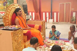 A man seated on an ornate gold-carved throne with red cushions, wearing an orange saffron robe and a long dark rudraksha mala, his feet resting on a gold-edged platform decorated with pink lotus flowers. A woman in a teal and blue sari kneels with palms joined in namaskar, and a bald man in white dhoti and shawl with prayer beads also sits with joined hands. Before the throne are gold trays holding lotus flowers, fruit, small oil lamps and brass bowls, with a tray of apples and bananas on a side table. Behind them are orange and red walls with white stripes, a microphone stand, and a gold-framed carved panel. Several seated devotees are partially visible in the foreground.
