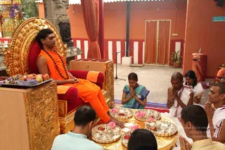 A photo of a padapuja ceremony in an open courtyard with red and white striped walls. A man seated on an ornate gold throne with red cushions wears orange robes, a rudraksha mala and a loose orange shawl, with his feet resting on a golden tray. Devotees kneel before him with palms pressed together: a woman in a teal and blue sari, a bald man in white dhoti and shawl, a man in white with folded hands, and a partly visible man in the foreground. Silver trays hold pink lotus flowers, red apples and small brass vessels. A microphone stand stands behind, with red curtains and a potted plant at right.