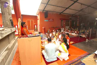 A wide photograph of an indoor satsang hall with orange-painted walls and a corrugated metal roof. On the left, a man in an orange robe with a garland sits on a raised golden-trimmed seat beside a carved stone pillar, with a wooden lectern and offering trays in front of him. Seated on red and orange mats below him, devotees in white and yellow clothing sit with hands pressed together in namaste, including a man in pale blue kurta and a woman in white with a yellow shawl. A stack of brass plates and offerings lies on the floor, and a video camera on a tripod films the gathering at the right, with more seated devotees in the background. Some devotees wear orange shawls.