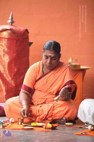 A woman in orange sari with maroon border sits cross-legged on a mat before a brass plate of yellow flowers and small brass vessels, holding a brass bell in her right hand and a small spoon in her left. She has a white tilak and red bindi, with a gold necklace. Behind her a red draped structure and a lit oil lamp stand against an orange wall. Another person in white clothes is partly visible at the right edge. Ritual puja setting with brass implements and oil lamp.