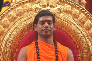 A man with closed eyes sits upright on an ornate gilded throne with a large carved golden back and a red cushion. He wears a saffron-orange draped garment over bare shoulders and a long rudraksha mala with dark beads hanging to his chest, with a red bindi on his forehead. The setting is a ceremonial seat in a temple or hall, and his expression is calm and meditative.