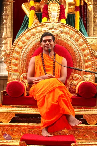A seated man in saffron-orange robes sits on an ornate gold throne with a deep red cushioned seat and red bolster pillows. He wears a dark rudraksha mala around his neck and a red bindi on his forehead, with hands resting in his lap and bare feet on a red footstool. A microphone on a stand is near him. Behind the throne hang yellow and magenta flower garlands and a golden kalasha-like finial. The setting suggests a satsang talk. a person is seated on the peetha, with saffron robes and rudraksha mala as the distinguishing cues.