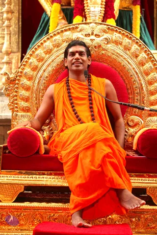 A man in an orange saffron garment sits on an ornate gold throne with red velvet cushions, speaking into a clip-on microphone. He wears a long dark rudraksha mala around his neck and a red bindi on his forehead, with bare feet resting on a red footstool. Behind him are yellow and red flower garlands draped over a golden structure and green drapes. a person: the saffron robe, rudraksha mala and ornate gold seating are consistent with the page's reference to a talk by Bhagavan a person, but the figure is seated and speaking rather than identified by face.