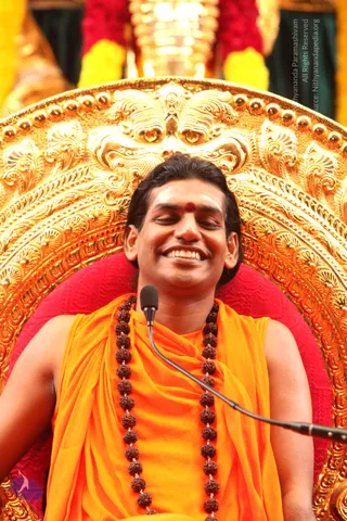 A smiling man with eyes closed sits on an ornate gold throne with a red cushion, backed by a gilded arch with floral and scroll carvings. He wears saffron-orange robes draped over his shoulders and a long rudraksha mala of brown beads, with a red bindi and white forehead markings. A microphone stands in front of him, and yellow and red garlands hang above. Swamiji (probable): the long swept-back hair, saffron clothing, rudraksha mala and throne match the page's claim, so he is treated as a probable in-person candidate. The image is a photograph of one seated person, with no other physical people visible.