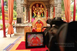 A photograph of a man seated on an ornate gold throne with red cushions, wearing orange saffron robes and a rudraksha mala, in front of a shrine with a deity murti of a seated couple in gold and orange garments decorated with garlands. A video camera is in the foreground, its monitor showing the same seated figure in orange. Red and orange curtains frame the carved stone pavilion, with a red carpet and a patterned rug below. One person is visible.